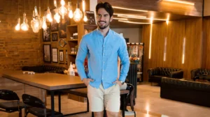 Um homem de camisa azul e short bege parado em uma barbearia, procurando outlet de roupas multimarcas.
